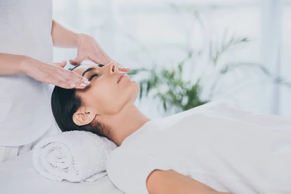 Cropped shot of young woman with closed eyes receiving reiki treatment on face — Stock Photo
