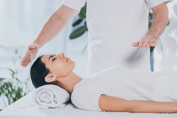 Tiro recortado de la mujer joven tranquila que recibe tratamiento reiki de curandero masculino - foto de stock