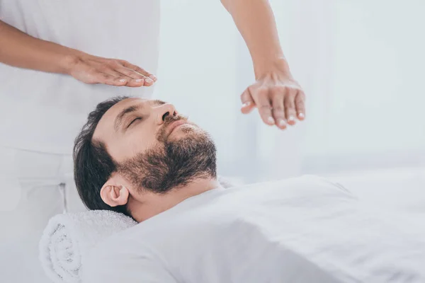 Hombre barbudo tranquilo acostado en la mesa de masaje y las manos de curandero haciendo sesión de tratamiento reiki — Stock Photo