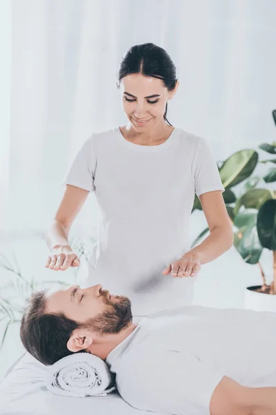 Sorrindo jovem curandeiro fazendo reiki sessão para acalmar homem barbudo com olhos fechados — Fotografia de Stock