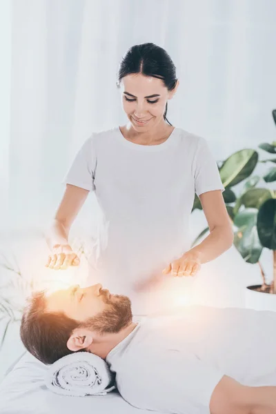 Sonriente joven curandera haciendo sesión de reiki para calmar al hombre barbudo con los ojos cerrados - foto de stock