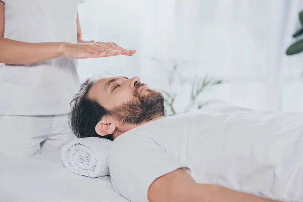 Recortado disparo de barbudo hombre con los ojos cerrados recibir tratamiento reiki por encima de la cabeza - foto de stock