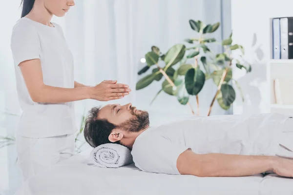 Vue latérale de l'homme barbu calme avec les yeux fermés recevant un traitement reiki sur la table de massage — Photo de stock