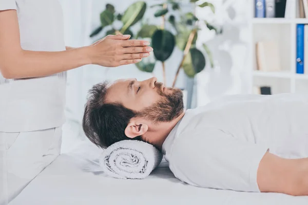 Vista lateral del hombre barbudo con los ojos cerrados recibiendo tratamiento reiki por encima de la cabeza — Stock Photo