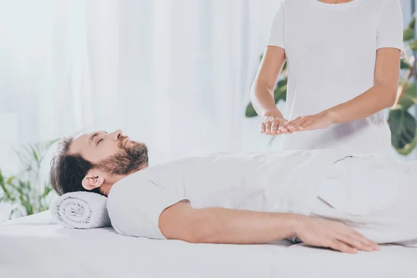 Tiro recortado de hombre barbudo tranquilo con los ojos cerrados acostado en la mesa de masaje y recibir tratamiento reiki - foto de stock