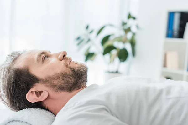 Seitenansicht eines bärtigen Mannes auf Massagetisch liegend und in Arztpraxis aufblickend — Stockfoto