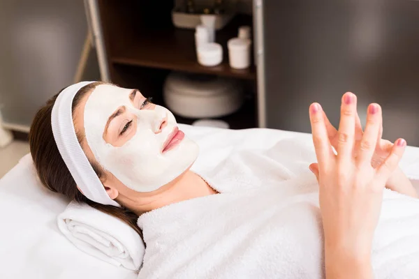 Frau liegt im weißen Bademantel mit aufgesetzter Gesichtsmaske im Schönheitssalon — Stockfoto