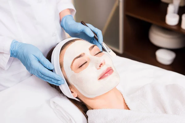 Esthéticienne debout et tenant brosse cosmétique près de la femme avec masque cosmétique sur le visage au salon de beauté — Photo de stock
