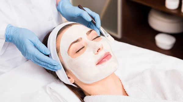 Esthéticienne debout près de la femme avec masque cosmétique sur le visage et tenant brosse cosmétique au salon de beauté — Photo de stock