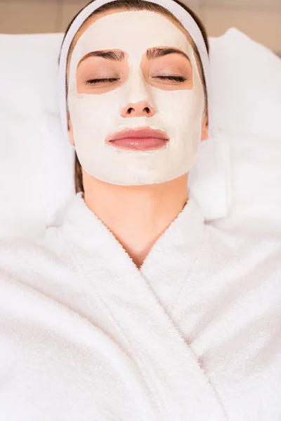 Vista superior de la mujer acostada en albornoz blanco con mascarilla aplicada en el salón de belleza - foto de stock