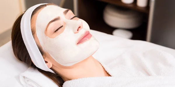 Woman lying in white bathrobe with applied facial mask and smiling at beauty salon — Stock Photo