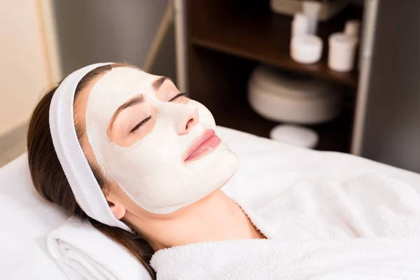 Mulher deitada em roupão de banho branco com máscara facial aplicada, sorrindo e olhos fechados no salão de beleza — Fotografia de Stock