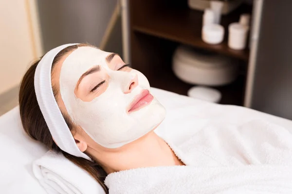 Mujer acostada en albornoz blanco con máscara facial aplicada, ojos sonrientes y cerrados en el salón de belleza - foto de stock