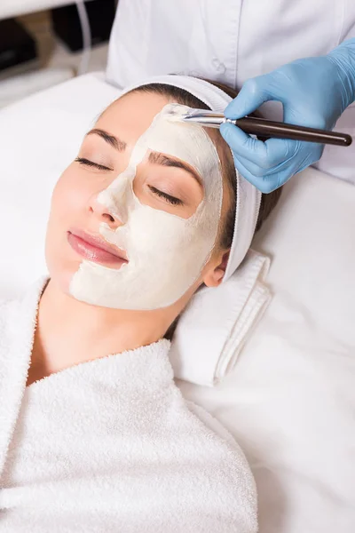 Beautician applying cosmetic mask on half woman face with cosmetic brush at beauty salon — Stock Photo