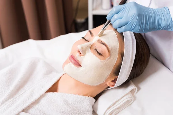 Estetista che applica la maschera cosmetica con spazzola cosmetica a donna su metà faccia a salone di bellezza — Foto stock