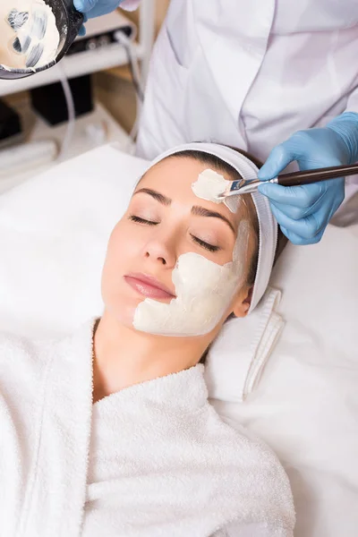 Beautician applying cosmetic mask on woman face with cosmetic brush and holding vessel at beauty salon — Stock Photo