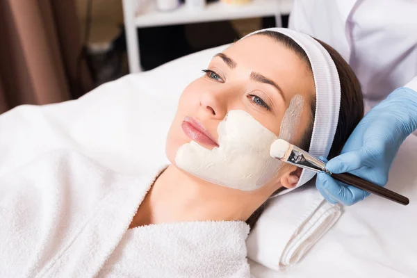 Esteticista aplicando máscara cosmética no rosto da mulher parte com escova cosmética no salão de beleza — Fotografia de Stock