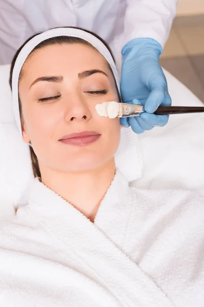Kosmetikerin beginnt Kosmetikmaske auf Gesicht der Frau mit Kosmetikpinsel im Schönheitssalon aufzutragen — Stockfoto