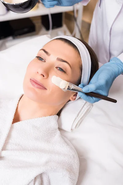 Esthéticienne commence à appliquer un masque cosmétique avec pinceau cosmétique sur le visage de la femme au salon de beauté — Photo de stock
