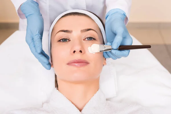 Kosmetikerin trägt Kosmetikmaske mit Kosmetikpinsel auf Gesicht zu Frau im Schönheitssalon auf — Stockfoto