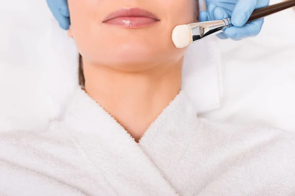 Cropped view of beautician applying cosmetic mask on woman face with cosmetic brush at beauty salon — Stock Photo