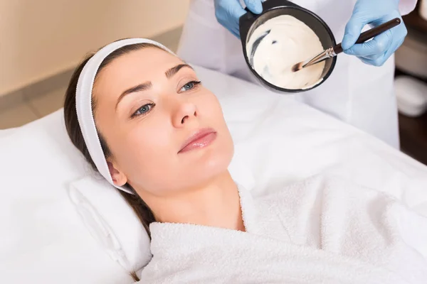 Séduisante femme couchée en peignoir et bandeau tandis que l'esthéticienne mélange masque facial au salon de beauté — Photo de stock