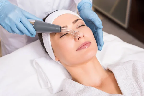 Beautician doing physiotherapy whith darsonvalization equipment on woman face at beauty salon — Stock Photo