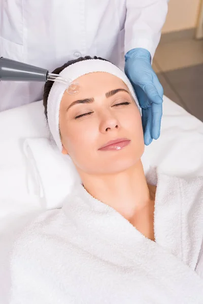 Beautician doing darsonvalization to woman on face at beauty salon — Stock Photo
