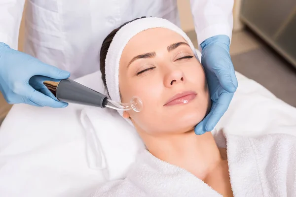 Beautician doing darsonvalization with equipment to woman on cheek at beauty salon — Stock Photo