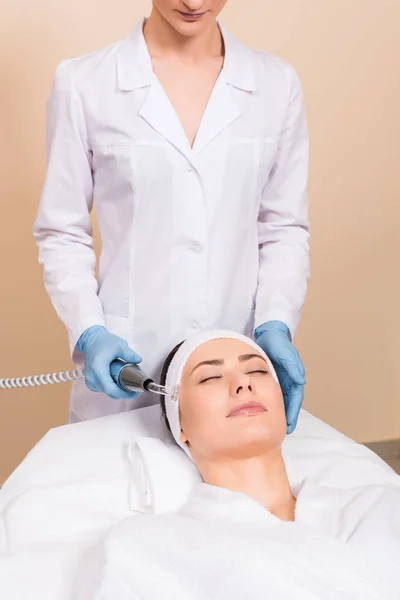Beautician doing darsonvalization with equipment to woman on face at beauty salon — Stock Photo