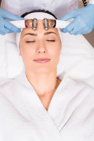 Esteticista fazendo massagem facial com equipamentos para mulher no salão de beleza — Fotografia de Stock