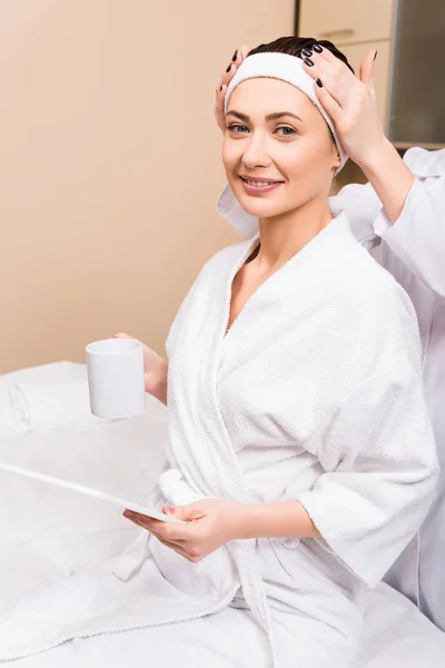 Woman sitting, holding cup and digital tablet while beautician correcting hairband at beauty salon — Stock Photo