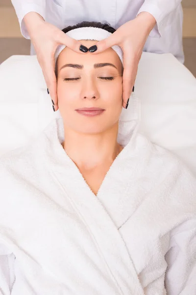 Cosmetólogo haciendo masaje facial manual a la mujer acostada en el albornoz y la banda de pelo en el salón de belleza - foto de stock