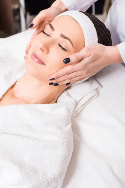 Esteticista dando masaje facial manual a la mujer acostada en el albornoz y la banda de pelo en el salón de belleza - foto de stock