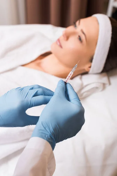 Selective focus of syringe in cosmetologist hands in front of woman at beauty salon — Stock Photo