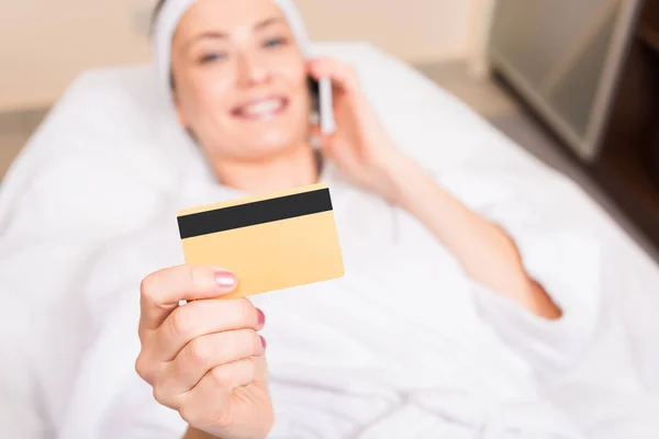 Mulher atraente deitado, falando no smartphone e segurando cartão de crédito no salão de beleza — Fotografia de Stock