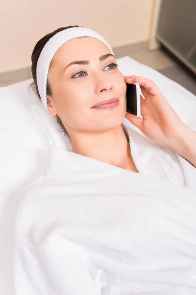 Attractive woman talking on smartphone and lying in white bathrobe at beauty salon — Stock Photo