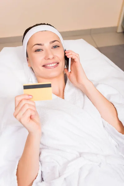 Attractive woman lying in white bathrobe, talking on smartphone and holding credit card at beauty salon — Stock Photo