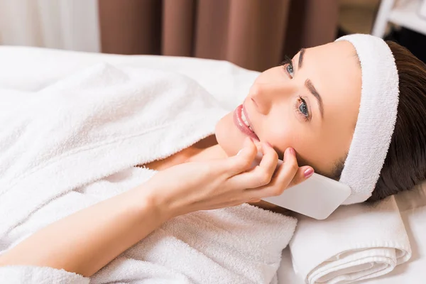 Attractive woman lying in white bathrobe and talking on smartphone at beauty salon — Stock Photo