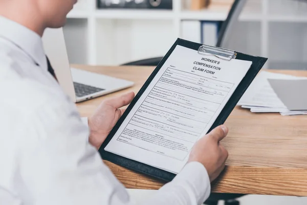 Visión parcial del empresario que lee el formulario de reclamación de compensación mientras está sentado en el escritorio de la oficina, concepto de compensación - foto de stock