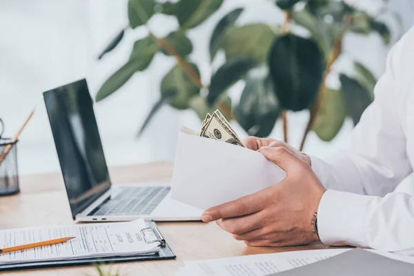 Visión parcial del empresario sosteniendo sobre con dinero en las manos en el lugar de trabajo, concepto de compensación - foto de stock