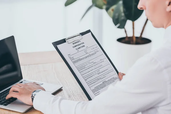 Vue recadrée de l'homme d'affaires tapant sur le clavier d'ordinateur portable tout en lisant la demande d'indemnisation sur le lieu de travail — Photo de stock