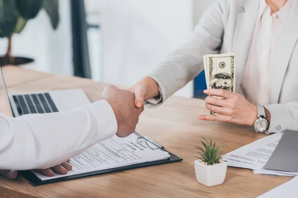 Abgeschnittene Ansicht einer Geschäftsfrau, die Mann die Hand schüttelt und am Arbeitsplatz Bargeld in der Hand hält, Vergütungskonzept — Stockfoto