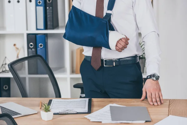 Обрізаний вид працівника з розбитою рукою в бинті, що стоїть біля столу в офісі, концепція компенсації — стокове фото