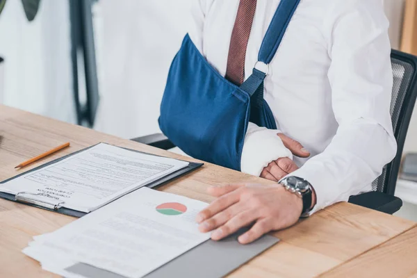 Vista recortada del trabajador con el brazo roto en el vendaje sentado en la mesa en la oficina, concepto de compensación - foto de stock