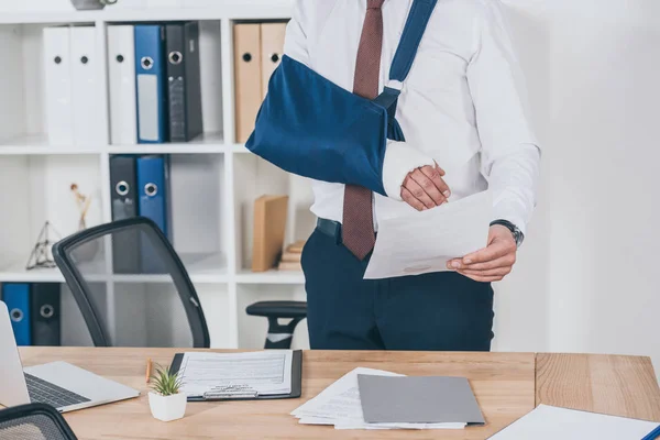 Обрізаний вид працівника з розбитою рукою в пов'язці стоїть і тримає папір над столом в офісі, концепція компенсації — стокове фото
