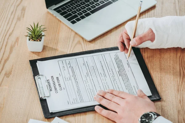 Cropped view of man with broken arm in gypsum signing compensation form on table in office, compensation concept — Stock Photo