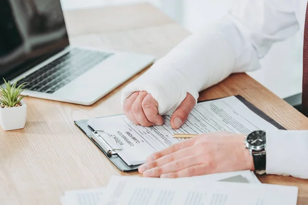 Vista recortada del trabajador con el brazo roto en yeso sentado a la mesa y cogido de la mano en forma de compensación en la oficina, concepto de compensación - foto de stock