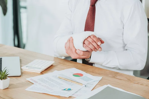 Ausgeschnittene Ansicht eines Arbeiters mit gebrochenem Arm, der im Büro an Dokumenten sitzt, Vergütungskonzept — Stockfoto