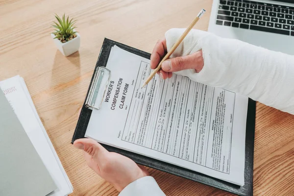 Cropped view of worker with broken arm in gypsum sitting at table and writing on compensation form in office, compensation concept — Stock Photo